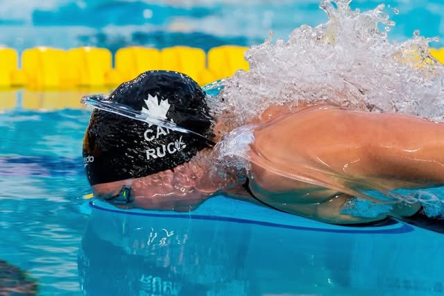 Taylor Ruck in a Team Canada cap and goggles breaking the surface of a pool lane