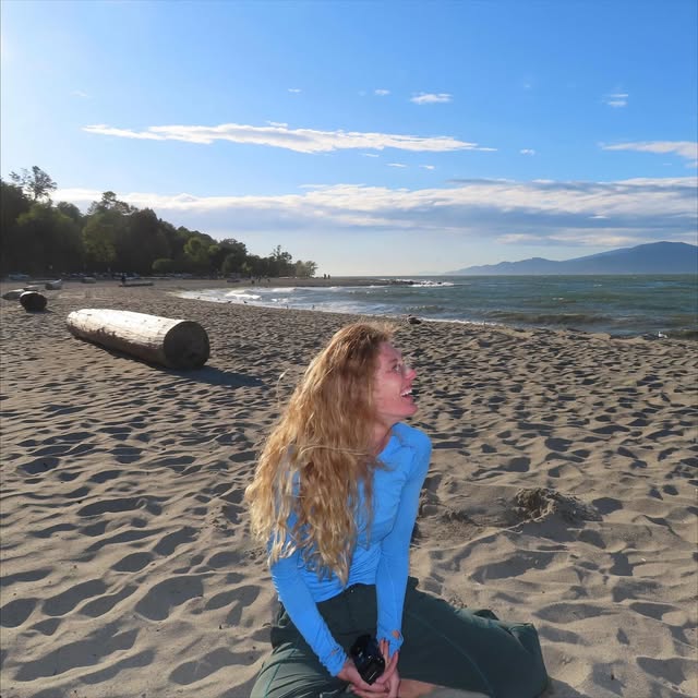 Taylor Ruck smiling on a sandy beach with driftwood, waves and distant mountains
